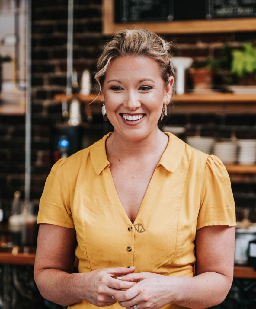 A smiling woman in a yellow blouse standing in a cozy cafe environment, exuding warmth and hospitality.