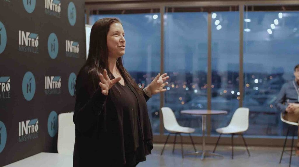 A professional woman engages in an animated conversation or presentation in a modern office setting with large windows overlooking the city during evening hours.