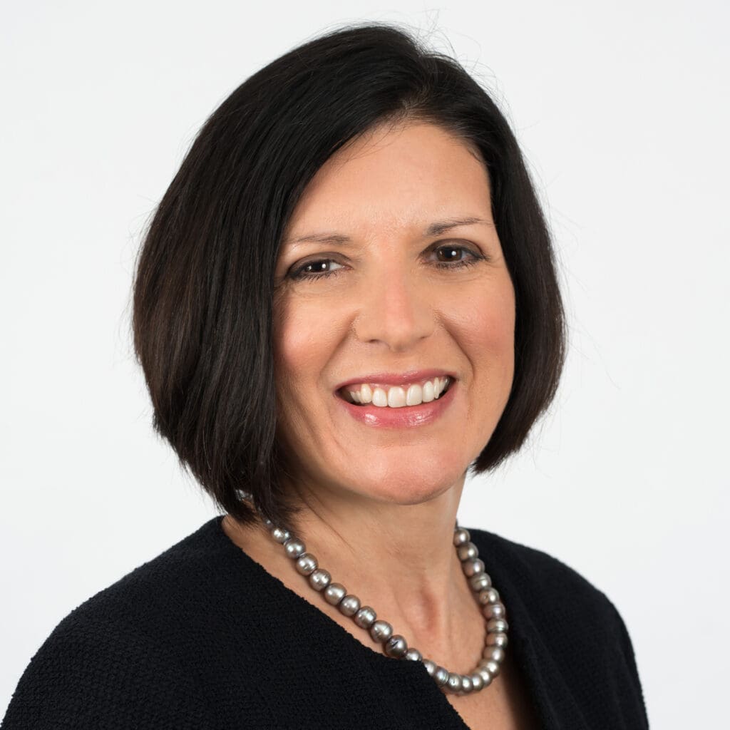 Professional woman with a confident smile wearing a black outfit and a pearl necklace against a white background.