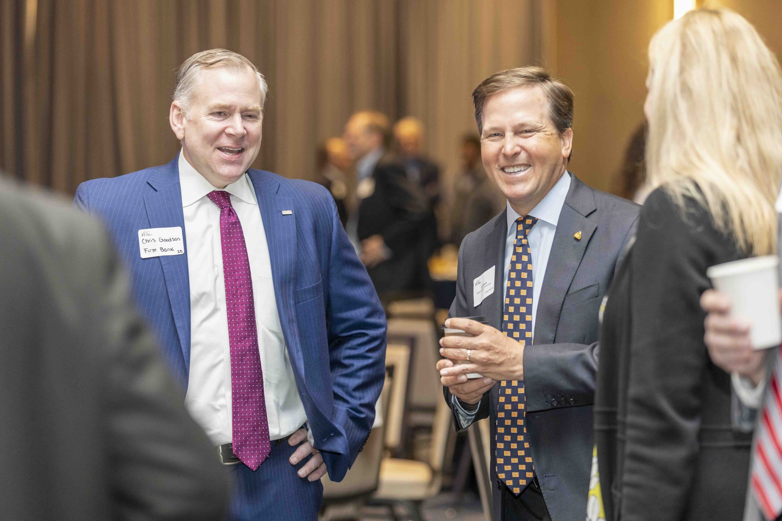 Two professionals engaged in a friendly conversation at a networking event, with others mingling in the background.