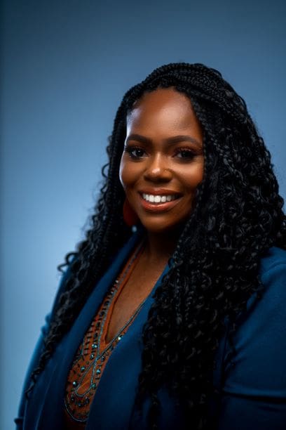 Confident professional woman with a bright smile, sporting long braided hair and a stylish blue blazer over an intricately patterned top, set against a soft blue background.