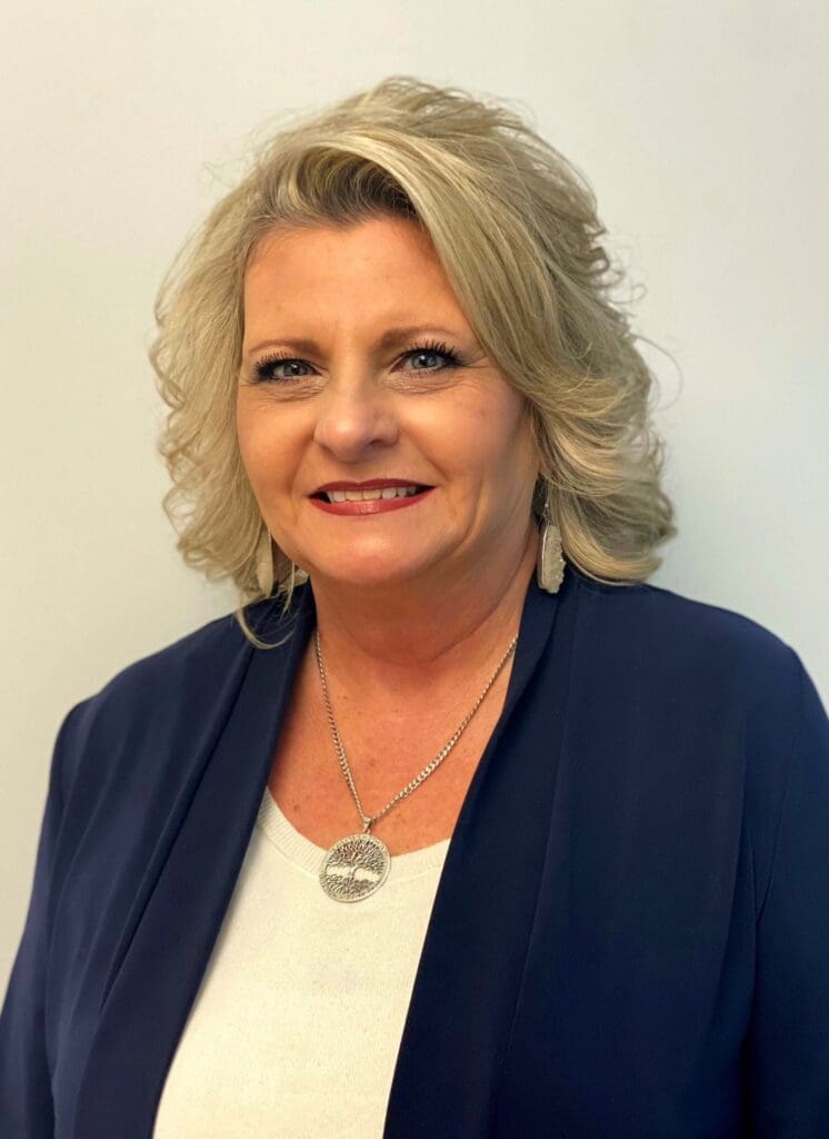 A professional woman with a friendly smile, wearing a navy blazer and a white top, complemented by a stylish necklace, poses for a portrait against a neutral background.