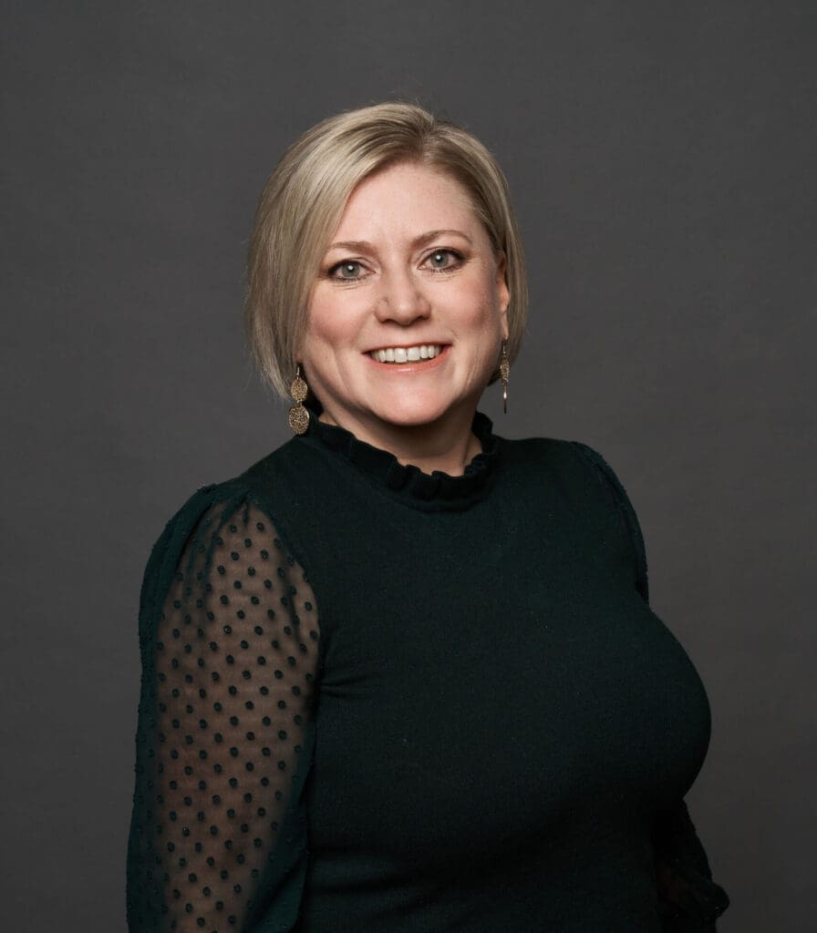 Confident woman with a warm smile, wearing a stylish black top with sheer dotted sleeves, poses for a professional portrait against a grey background.