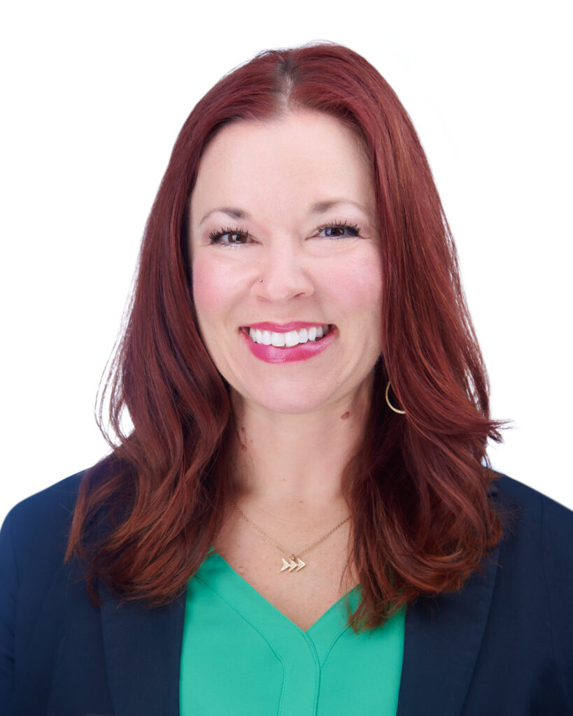 A professional headshot of a smiling woman with red hair wearing a teal top and a dark blazer against a white background.