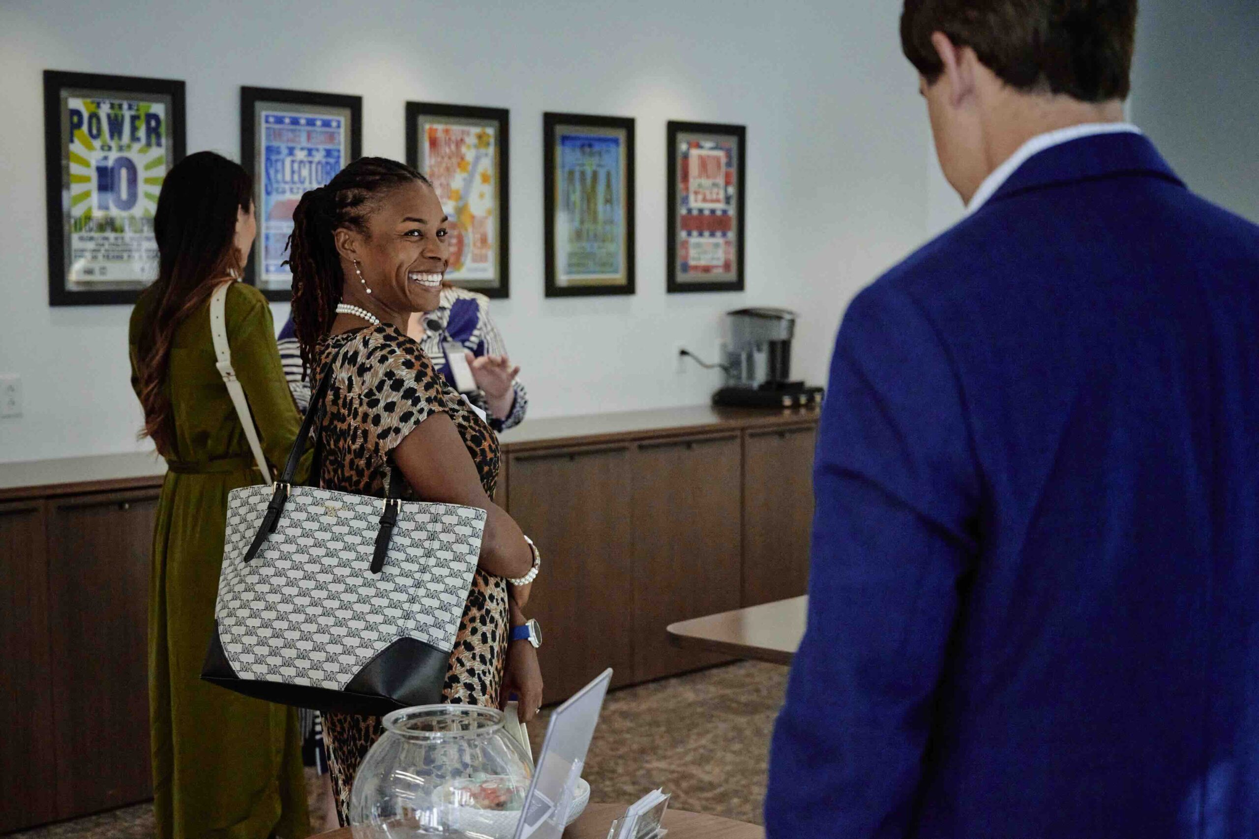 A professional encounter: a woman with a cheerful expression holding a designer tote bag exchanges a conversation with a man in a blue blazer in an office environment adorned with colorful posters on the wall.