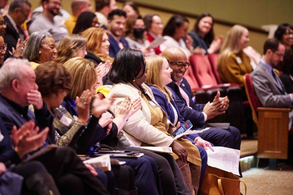 An engaged audience clapping during a conference or event, with expressions of interest and enjoyment on their faces.