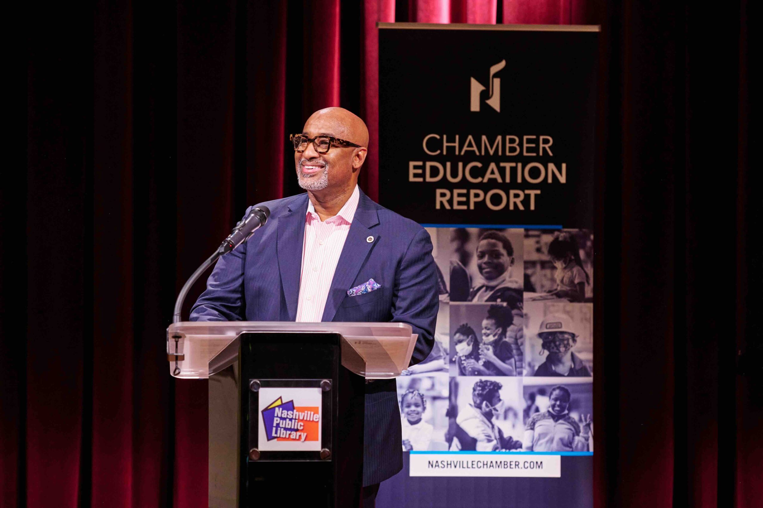 A confident speaker addressing an audience from a podium at an event focused on education, hosted by the nashville chamber, as indicated by the banner beside him.