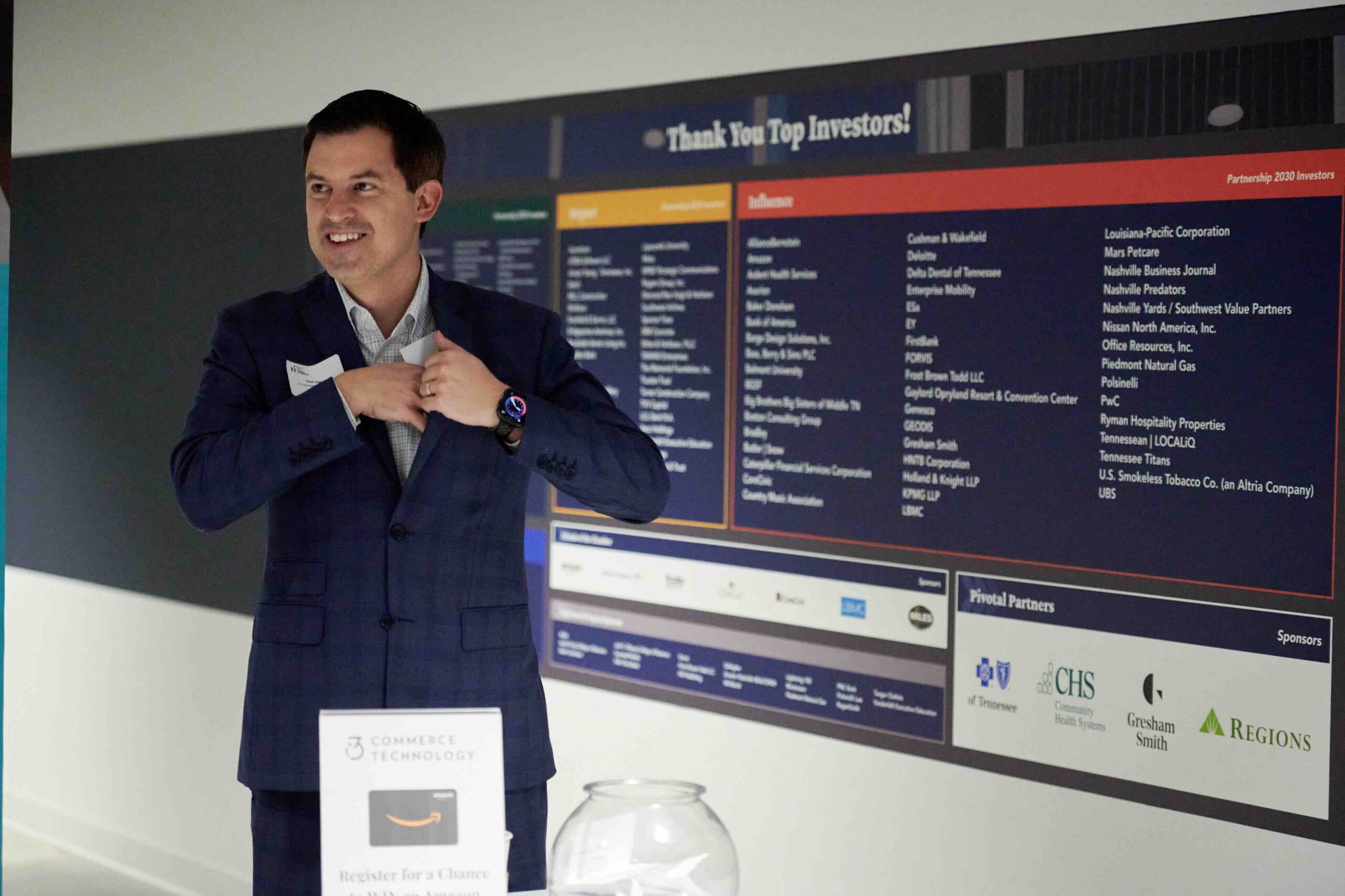 A man in a dark suit and bow tie speaking with a smile, gesturing with his hand, standing next to a sign thanking top investors at a formal event or presentation.