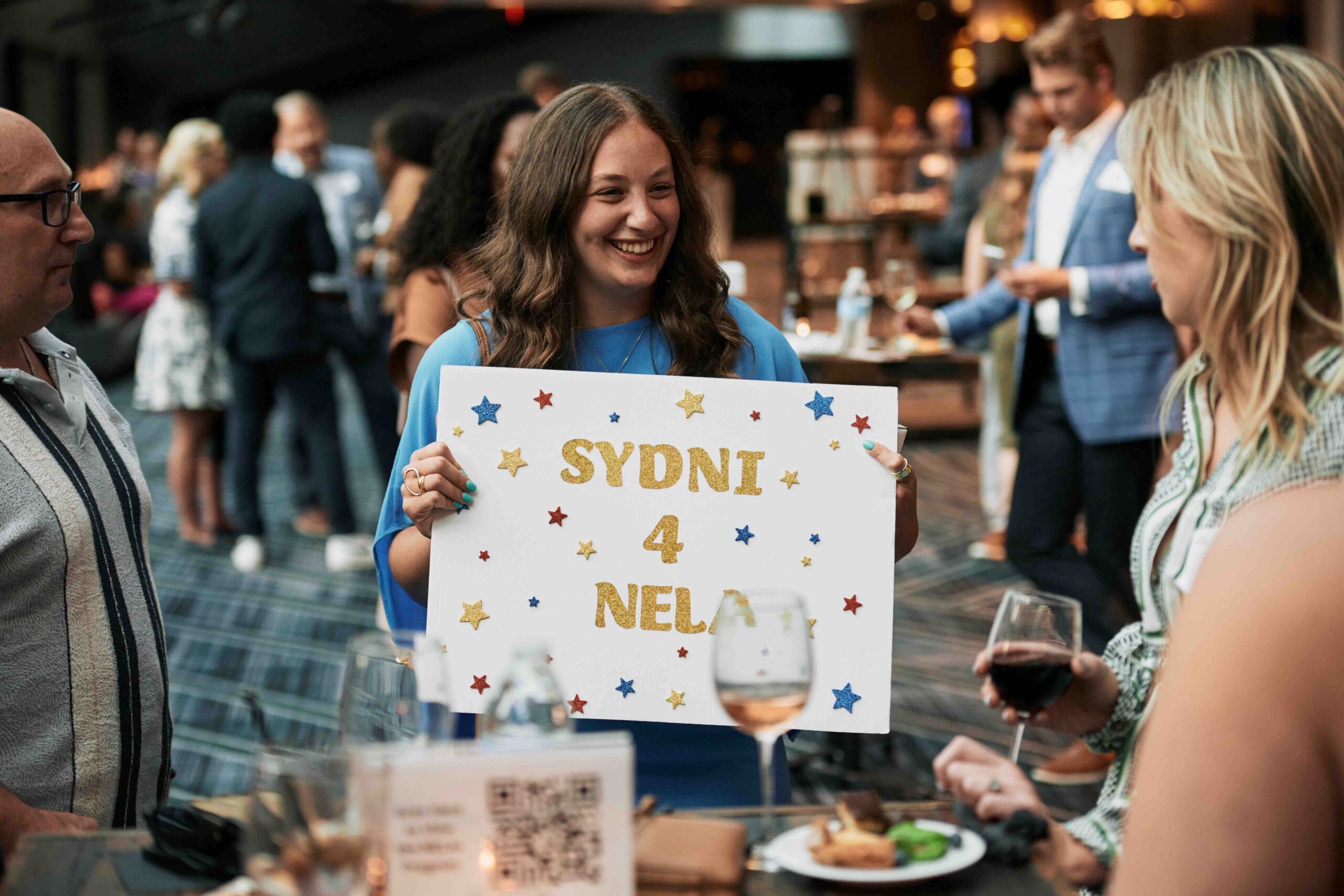 A joyful gathering with a smiling woman holding a custom sign that reads "sydni 4 nela," surrounded by guests enjoying drinks and conversation at a social event.